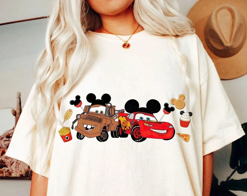 Puede incluir: Camiseta color crema con un gr&aacute;fico de dos coches de dibujos animados, uno marr&oacute;n y otro rojo, con orejas de Mickey Mouse. El dise&ntilde;o incluye iconos de comida como patatas fritas, un gofre y una magdalena. La camiseta es de estilo casual.