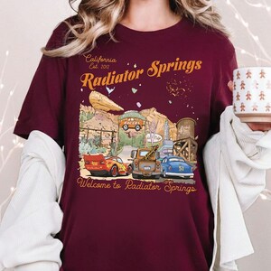 May include: Burgundy t-shirt featuring a colorful graphic of the town of Radiator Springs from the movie Cars. The design includes the text "California Est. 2012" and "Welcome to Radiator Springs". The graphic includes several cars and buildings.