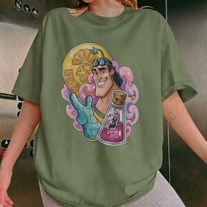 May include: Olive green t-shirt featuring a cartoon illustration. The graphic depicts a smiling character holding a gold coin and a potion bottle with a cork stopper. The design includes pink and purple accents.