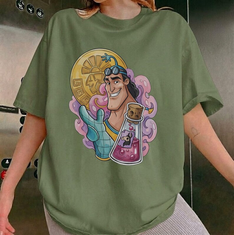 May include: Olive green t-shirt featuring a cartoon illustration. The graphic depicts a smiling character holding a gold coin and a potion bottle with a cork stopper. The design includes pink and purple accents.