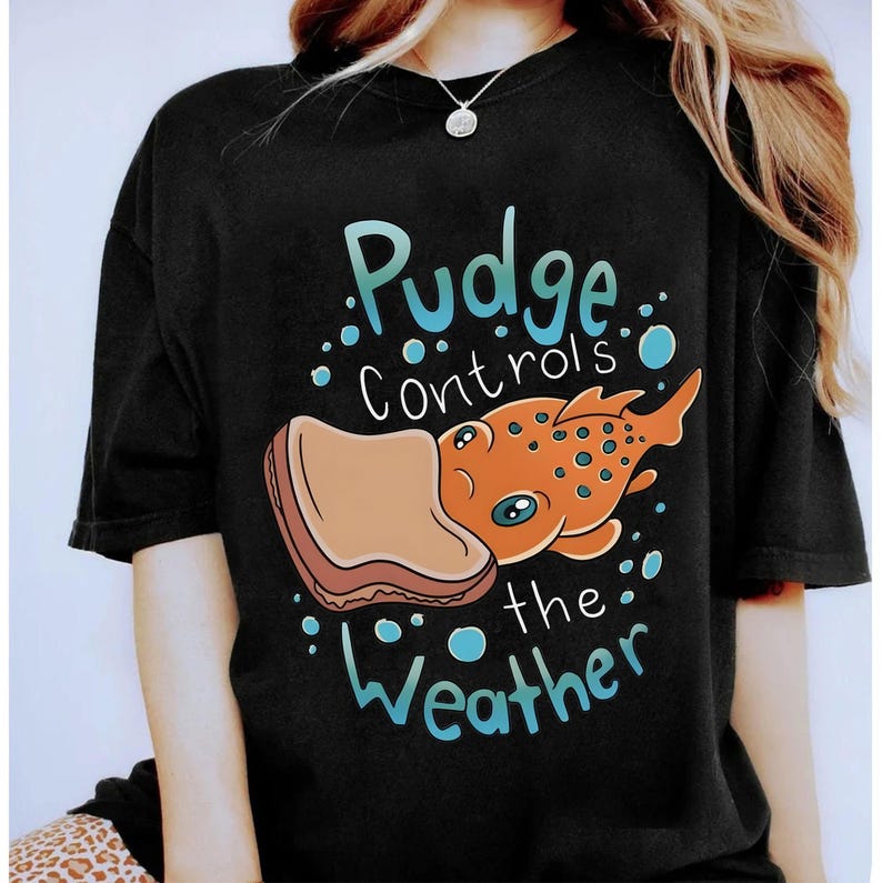 May include: Black t-shirt featuring a cartoon design. The graphic includes an orange fish with blue spots and a piece of toast, with the text "Pudge Controls the Weather" in blue and white.