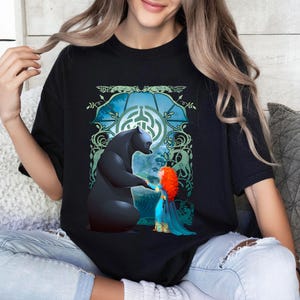 Op de afbeelding: Zwart T-shirt met een grafische print van een beer en een meisje met rood haar. De beer steekt een poot uit naar het meisje. De grafiek is omgeven door groene wijnstokken en een blauwe cirkel met een Keltisch knoopontwerp.