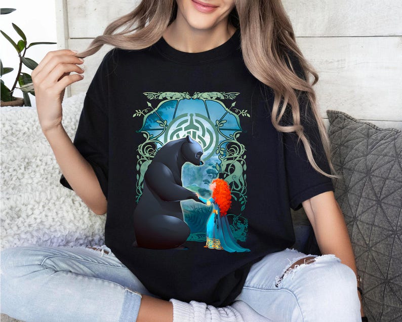 Op de afbeelding: Zwart T-shirt met een grafische print van een beer en een meisje met rood haar. De beer steekt een poot uit naar het meisje. De grafiek is omgeven door groene wijnstokken en een blauwe cirkel met een Keltisch knoopontwerp.