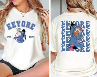 Può includere: T-shirt color crema con Eeyore di Winnie the Pooh. La parte anteriore mostra "Eeyore" in lettere blu a blocchi sopra una grafica circolare di Eeyore. Il retro mostra Eeyore che abbraccia un orsacchiotto, con il nome ripetuto.