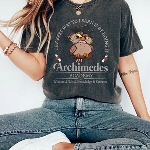 May include: Dark gray t-shirt featuring a circular design with an owl wearing a graduation cap, the text "The best way to learn is by doing it," and "Archimedes Academy." The shirt also includes the text "Wisdom & Wit & Knowledge & Sarcasm."