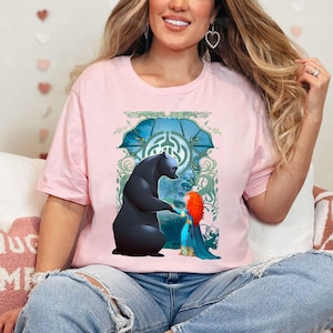 Op de afbeelding: Een roze T-shirt met een grafische print van een zwarte beer en een meisje met rood haar. De beer en het meisje staan ​​naast elkaar in een bosrijke omgeving. De afbeelding is omgeven door een groen en blauw ontwerp.