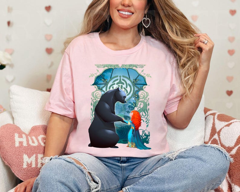 Op de afbeelding: Een roze T-shirt met een grafische print van een zwarte beer en een meisje met rood haar. De beer en het meisje staan ​​naast elkaar in een bosrijke omgeving. De afbeelding is omgeven door een groen en blauw ontwerp.