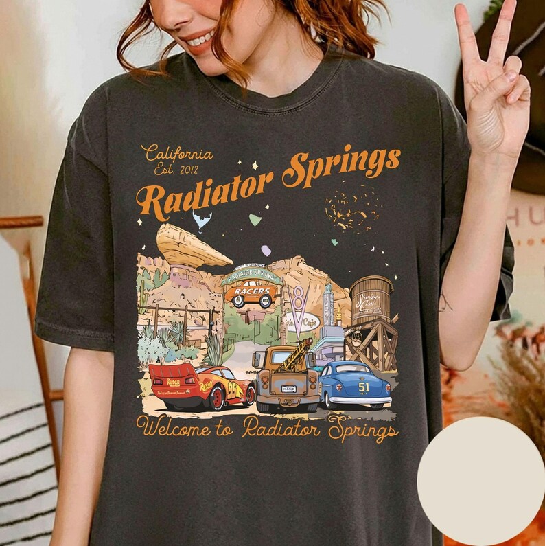May include: Dark gray t-shirt featuring a graphic of Radiator Springs, with the text "California Est. 2012 Radiator Springs" in orange. The design includes cartoon cars, a water tower, and desert scenery. The phrase "Welcome to Radiator Springs" is at the bottom.
