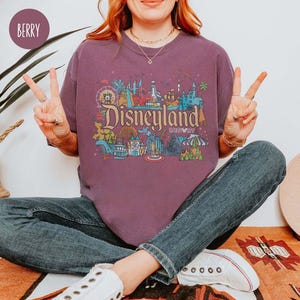 May include: A purple t-shirt with a graphic print of the Disneyland Resort logo and various attractions. The logo reads "Disneyland Resort".