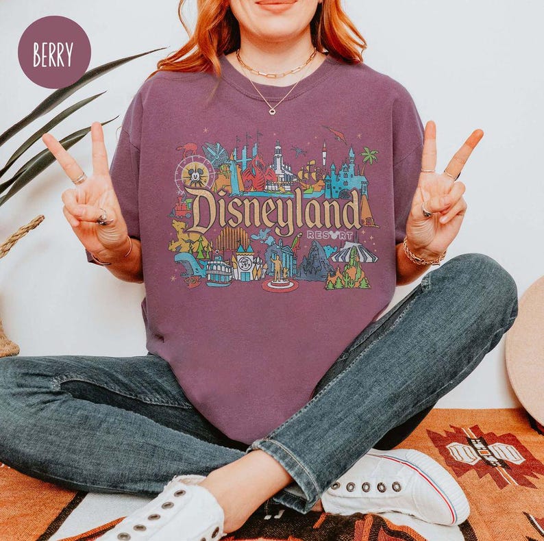 May include: A purple t-shirt with a graphic print of the Disneyland Resort logo and various attractions. The logo reads "Disneyland Resort".