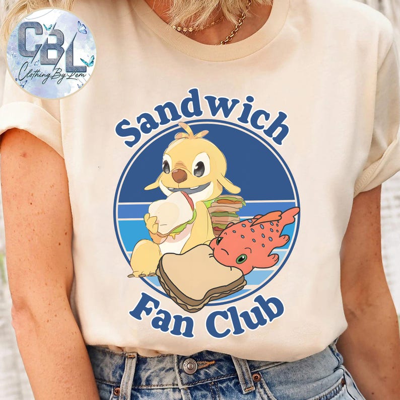 May include: A cream-coloured t-shirt with a cartoon character holding a sandwich, a red fish, and a slice of bread. The text "Sandwich Fan Club" is in a blue circle. The shirt has short sleeves and is made of a soft material.