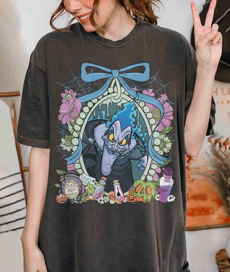 Pu&ograve; includere: T-shirt grigio scuro con un'illustrazione a cartoni animati di Ade di Hercules della Disney. Il design include Ade con fiamme blu, fiori, un fiocco blu e il testo "Deadly Night Shade".