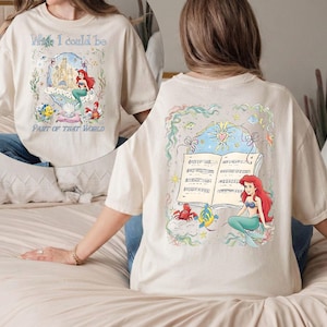 May include: Cream-colored t-shirt featuring a colorful illustration of a mermaid, a castle, and the text "Wish I could be Part of That World." The back of the shirt has a similar design with musical notes and a mermaid.