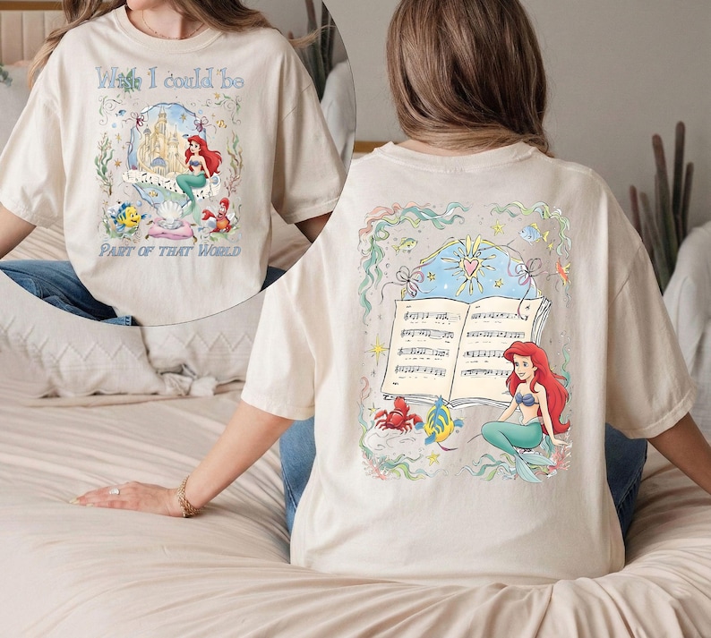 May include: Cream-colored t-shirt featuring a colorful illustration of a mermaid, a castle, and the text "Wish I could be Part of That World." The back of the shirt has a similar design with musical notes and a mermaid.