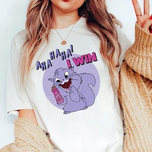 Puede incluir: Camiseta blanca con un dibujo de una ardilla de dibujos animados sosteniendo un objeto rosa, con el texto "AHA HAHA! I WIN" en un círculo morado. La ardilla es morada y sonríe. La camiseta es de estilo casual.