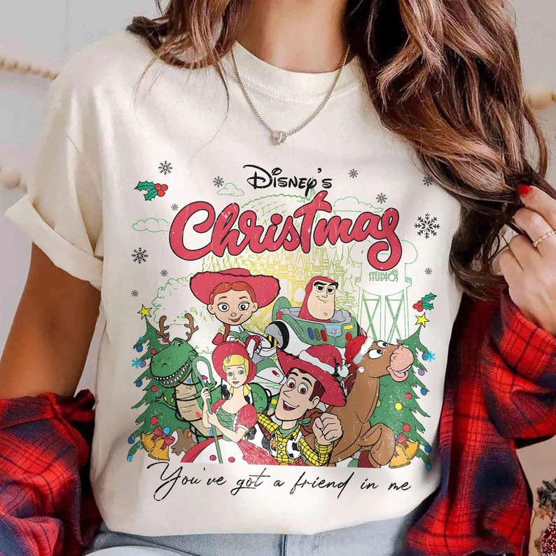 Może przedstawiać: Kremowy t-shirt z grafiką Disney's Christmas z postaciami z Toy Story. Projekt zawiera Woody'ego, Buzz'a Astrala, Jessie i inne postacie, a także frazę "You've got a friend in me."