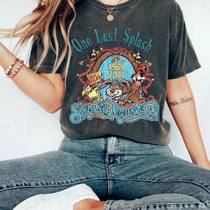 以下が含まれることがあります： 「One Last Splash」と「Splash Mountain」の文字が青色でプリントされた濃いグレーのTシャツ。漫画のキャラクターと1989年と2023年のグラフィックが特徴です。青いジーンズと白いスニーカーと合わせて着用されています。