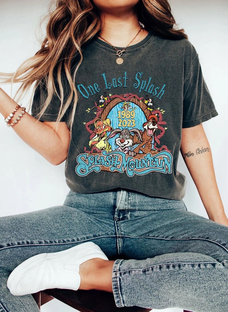 以下が含まれることがあります： 「One Last Splash」と「Splash Mountain」の文字が青色でプリントされた濃いグレーのTシャツ。漫画のキャラクターと1989年と2023年のグラフィックが特徴です。青いジーンズと白いスニーカーと合わせて着用されています。