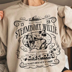 Puede incluir: Sudadera beige con un gráfico negro de Mickey Mouse al timón de un barco de vapor. El diseño incluye el texto "Steamboat Willie", "Mickey Mouse" y "Disney Cartoons". La sudadera tiene mangas largas y cuello redondo.