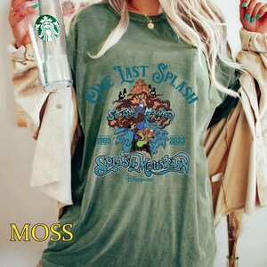 May include: Moss green t-shirt with the text "One Last Splash" and "Splash Mountain" in a teal font. The graphic features cartoon characters and the years 1989 and 2023. The shirt is a relaxed fit.