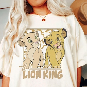 May include: Cream-colored t-shirt featuring a graphic of two lion cubs from The Lion King. The cubs are in a rectangular frame with a textured background. The words "The Lion King" are printed in a brown font below the image.