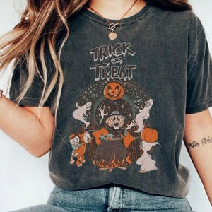 May include: Dark gray t-shirt with a vintage Halloween graphic. The design features the words "Trick or Treat" above a witch, ghosts, pumpkins, and other cartoon characters. The shirt has a slightly faded, worn look.