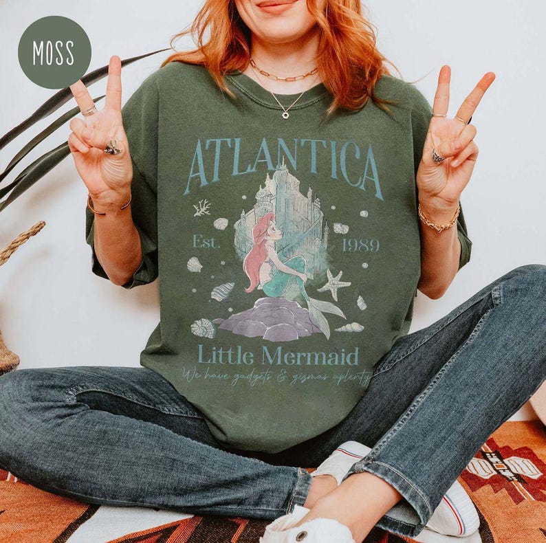 May include: A green t-shirt with a graphic of Ariel from The Little Mermaid in front of a castle. The text "Atlantica Est. 1989" and "Little Mermaid" is printed on the shirt.