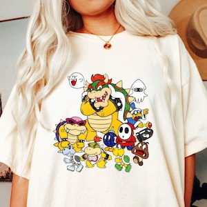 May include: Cream-colored t-shirt featuring a colorful graphic of various cartoon characters, including a large yellow Bowser, a white ghost, and other recognizable figures. The design is a vibrant mix of colors and cartoon styles.