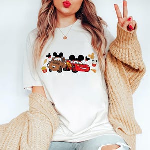 Puede incluir: Camiseta blanca con un gr&aacute;fico de dibujos animados de dos coches con orejas de Mickey Mouse e iconos de comida. Los coches son una gr&uacute;a marr&oacute;n y un coche de carreras rojo. La camiseta tiene cuello redondo y mangas cortas.