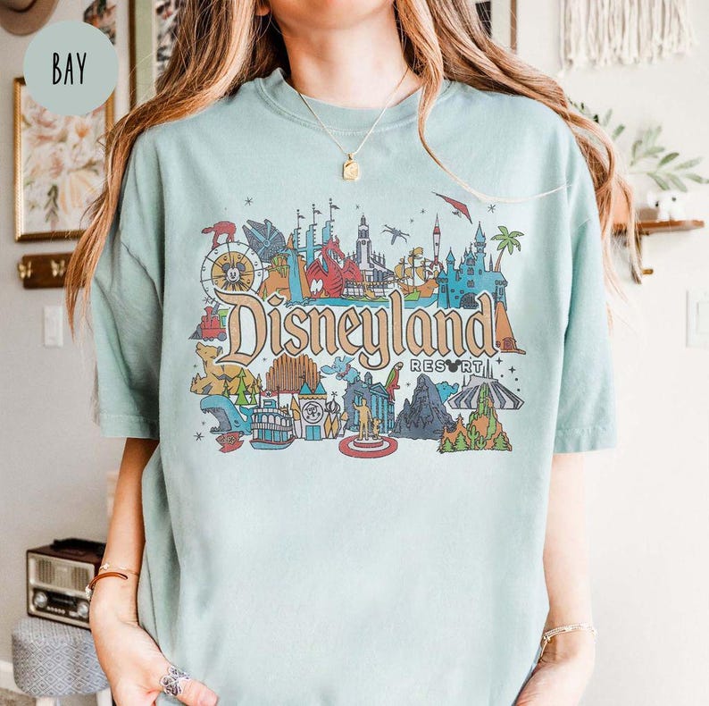 May include: A light blue t-shirt with a graphic print of Disneyland Resort. The graphic features a colorful illustration of various Disneyland attractions, including Sleeping Beauty Castle, the Matterhorn, and the Haunted Mansion.