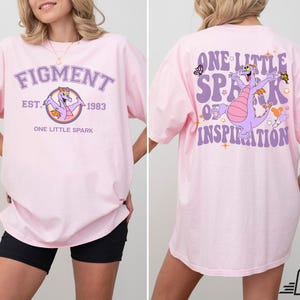 Op de afbeelding: Roze T-shirt met een paarse draak afbeelding en de tekst "Figment Est. 1983 One Little Spark" op de voorkant en "One Little Spark of Inspiration" op de achterkant.