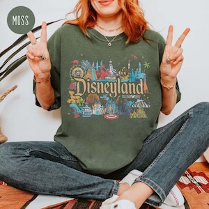 May include: A green t-shirt with a colorful graphic of Disneyland Resort. The graphic features the iconic Sleeping Beauty Castle and other attractions.