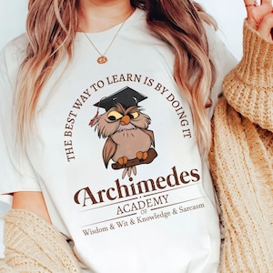 May include: White t-shirt featuring a cartoon owl wearing a graduation cap, perched on a branch. The text reads "THE BEST WAY TO LEARN IS BY DOING IT" and "Archimedes ACADEMY of Wisdom & Wit & Knowledge & Sarcasm."