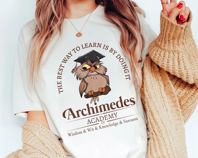 May include: White t-shirt featuring a cartoon owl wearing a graduation cap, perched on a branch. The text reads "THE BEST WAY TO LEARN IS BY DOING IT" and "Archimedes ACADEMY of Wisdom & Wit & Knowledge & Sarcasm."