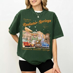 May include: Dark green t-shirt featuring a graphic of Radiator Springs, with the text "California Est. 2012 Radiator Springs" in orange. The graphic includes cartoon cars and desert scenery. The text "Welcome to Radiator Springs" is at the bottom.