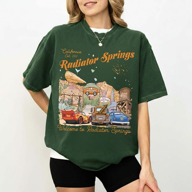 May include: Dark green t-shirt featuring a graphic of Radiator Springs, with the text "California Est. 2012 Radiator Springs" in orange. The graphic includes cartoon cars and desert scenery. The text "Welcome to Radiator Springs" is at the bottom.