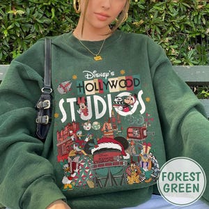 May include: Forest green sweatshirt featuring a colorful graphic of Disney's Hollywood Studios. The design includes Mickey and Minnie Mouse, Donald Duck, and other characters. The text "Disney's Hollywood Studios" is prominently displayed.