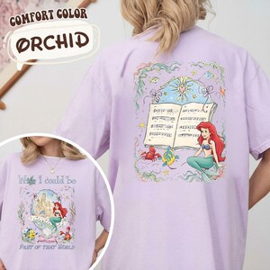 May include: Orchid-colored t-shirt featuring a graphic of Ariel from The Little Mermaid. The design includes a musical score, a crab, a fish, and Ariel. The shirt also has the text "Wish I could be Part of That World."