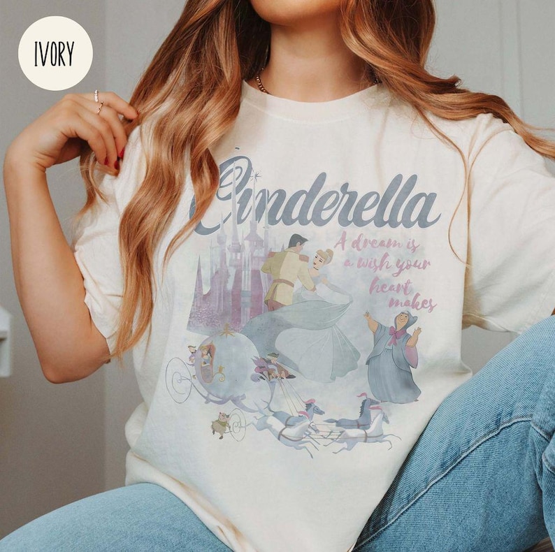May include: A cream coloured t-shirt featuring a vintage illustration of Cinderella and Prince Charming. The text "Cinderella" is in a large, stylised font. The text "A dream is a wish your heart makes" is printed below the illustration.