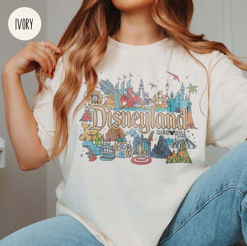 May include: A white t-shirt with a colorful graphic of Disneyland Resort. The graphic includes iconic landmarks and attractions, such as Sleeping Beauty Castle, Space Mountain, and the Haunted Mansion. The text "Disneyland Resort" is printed in a bold, black font.