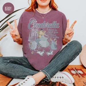May include: A purple t-shirt featuring a graphic of Cinderella and Prince Charming dancing. The text "Cinderella" is in a large, stylised font above the image. The text "A dream is a wish your heart makes" is below the image.