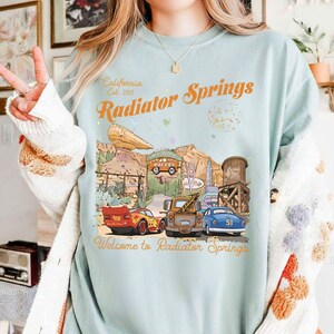 May include: Light blue t-shirt featuring a graphic of Radiator Springs from the movie Cars. The design includes the text "California Est. 2012" and "Radiator Springs" in orange, with images of the town and cars.