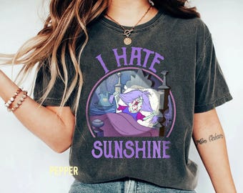 Può includere: T-shirt grigio scuro con la scritta "I HATE SUNSHINE" in viola. La grafica presenta un personaggio dei cartoni animati a letto, circondato da una cornice circolare viola. La maglietta è in stile casual e confortevole.