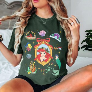 May include: A green t-shirt with a festive Christmas-themed design featuring Ariel from The Little Mermaid. The design includes a portrait of Ariel, seashells, a crown, and other nautical elements.