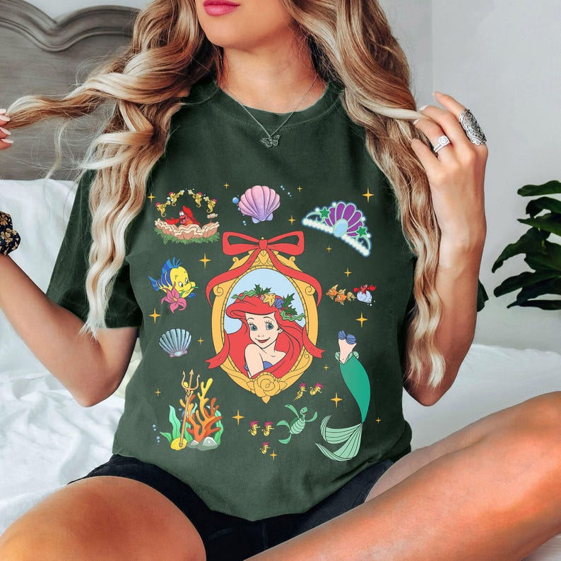 May include: A green t-shirt with a festive Christmas-themed design featuring Ariel from The Little Mermaid. The design includes a portrait of Ariel, seashells, a crown, and other nautical elements.