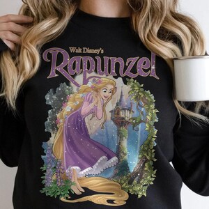 May include: Black sweatshirt featuring a graphic of Rapunzel from Walt Disney's animated film. The design includes Rapunzel in a purple dress with long blonde hair, a castle, and the word "Rapunzel" in a purple script.