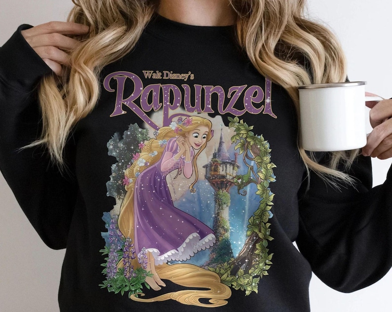 May include: Black sweatshirt featuring a graphic of Rapunzel from Walt Disney's animated film. The design includes Rapunzel in a purple dress with long blonde hair, a castle, and the word "Rapunzel" in a purple script.