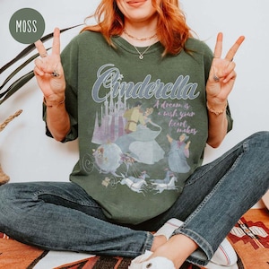 May include: A green t-shirt featuring a vintage-style graphic of Cinderella and Prince Charming. The text on the shirt reads "Cinderella, A dream is a wish your heart makes".