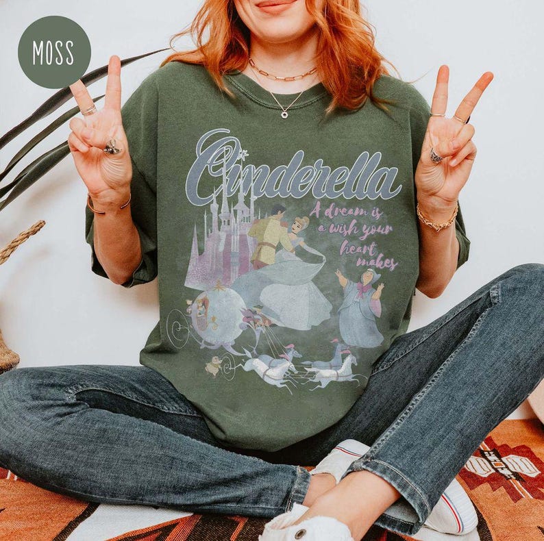 May include: A green t-shirt featuring a vintage-style graphic of Cinderella and Prince Charming. The text on the shirt reads "Cinderella, A dream is a wish your heart makes".