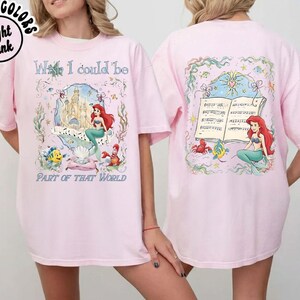 May include: Light pink t-shirt featuring a colorful graphic of a mermaid, castle, and text that reads "What I could be part of that world." The back of the shirt has a matching graphic of the mermaid and musical notes.